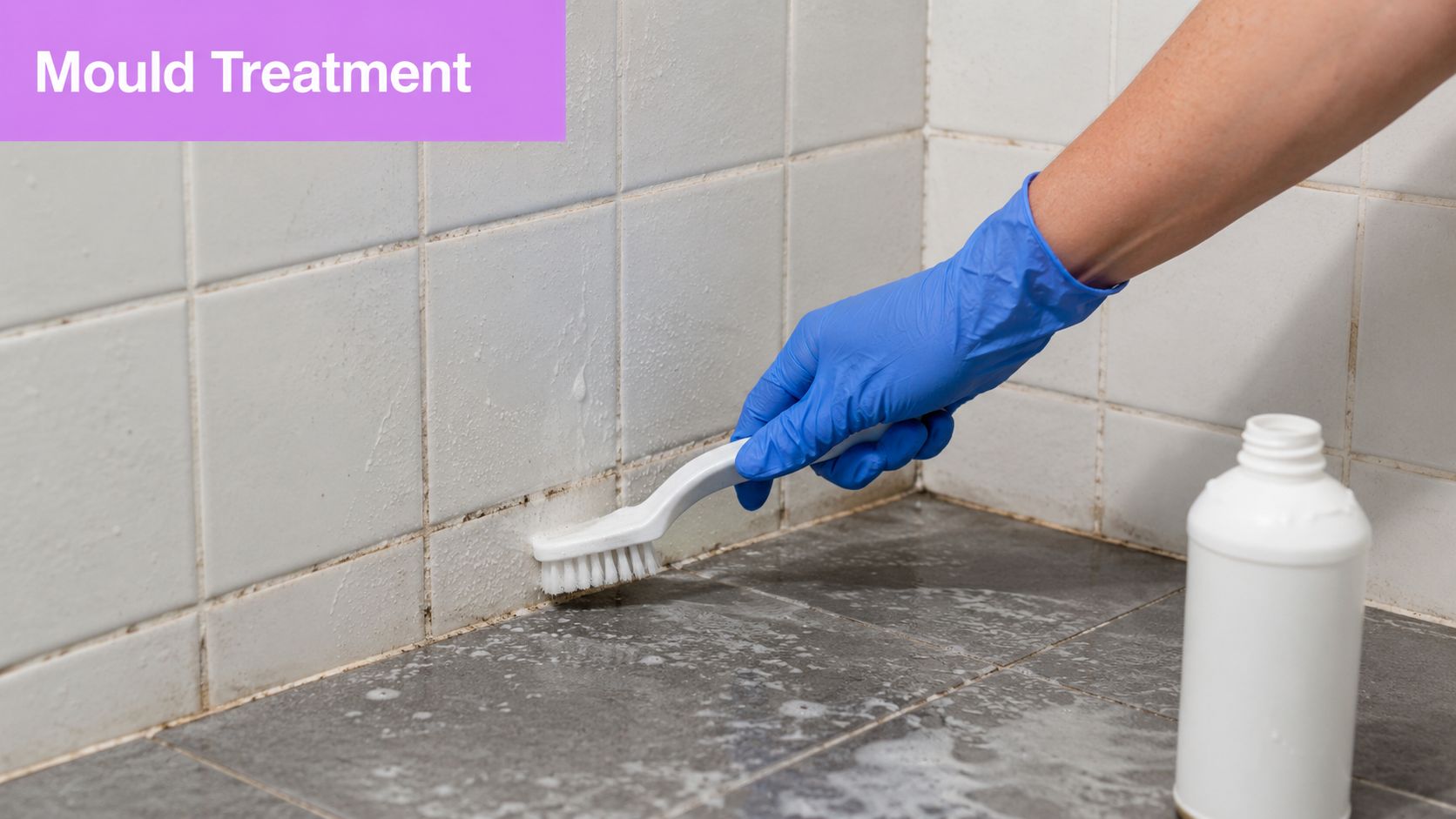 A person wearing blue rubber gloves scrubbing mould from bathroom tiles with a white plastic brush.