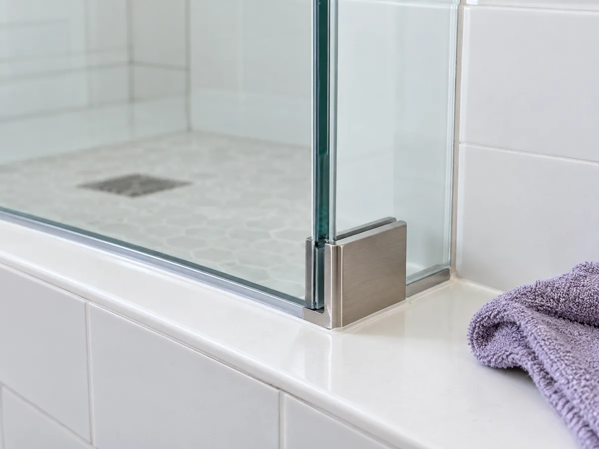 Lower glass shower edge meeting white tiles with an intact silicone seal and metal bracket